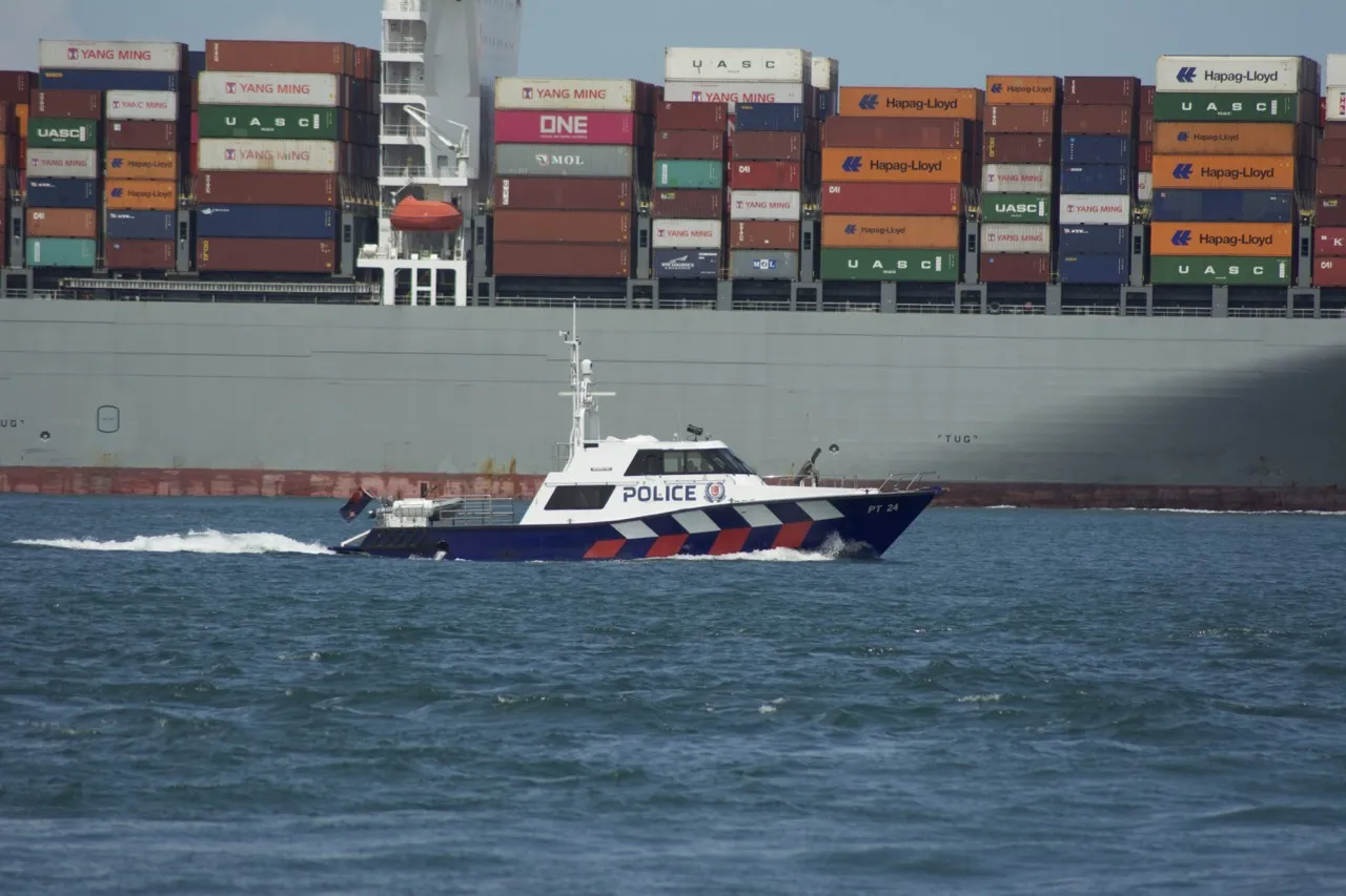 A blue and white police patrol boat moves fast past the side of a large container ship loaded with containers.