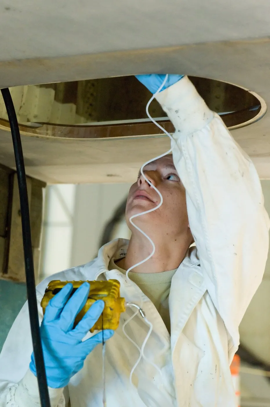 A person in white overalls and blue gloves reaches up into a round access hole in a tank with a sampling tube connected to a yellow handheld gas detector.