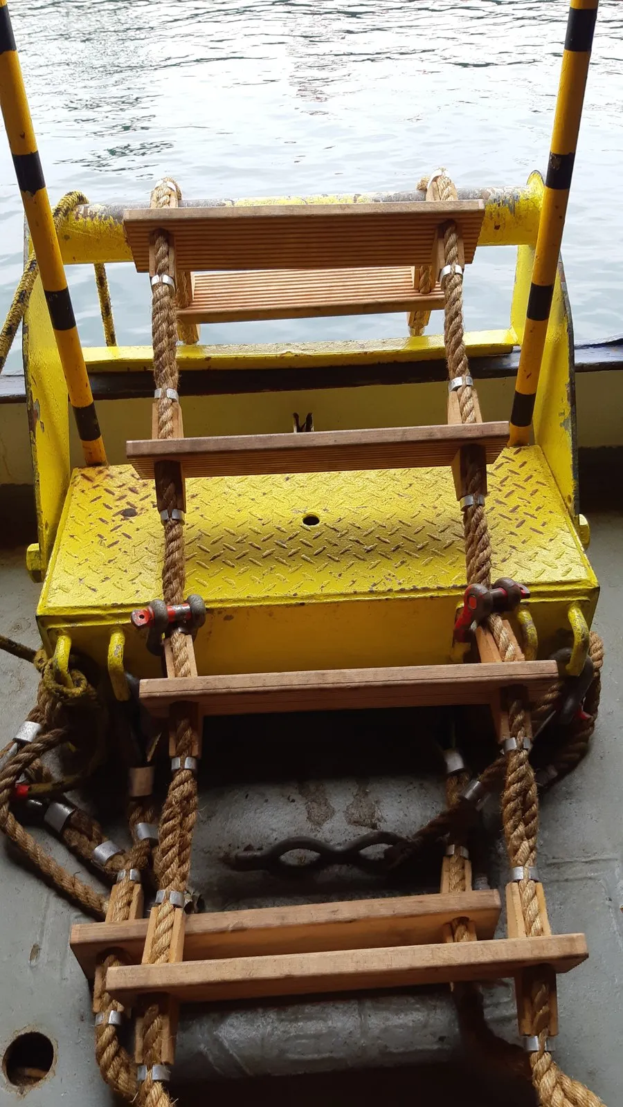 A rope pilot ladder with wooden steps hangs from a yellow steel platform at the side of a ship's deck.
