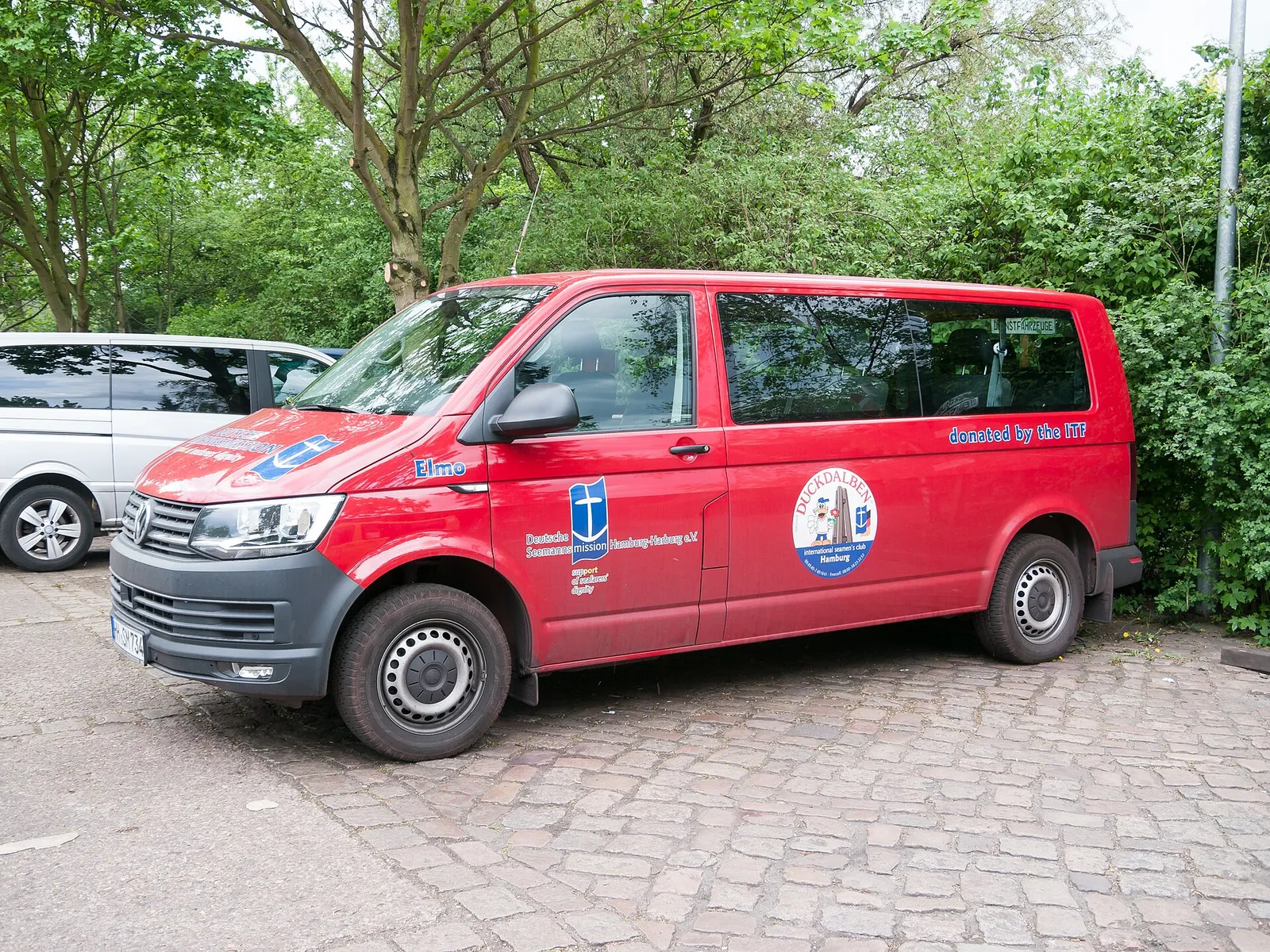 A red minibus parked on a cobbled street near green trees. It has the Duckdalben seafarers' club logo on the side.