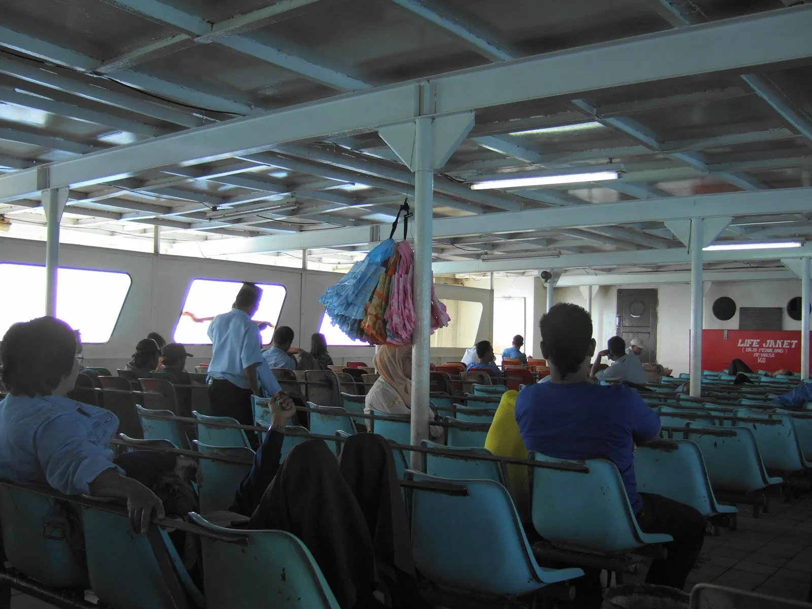 Rows of plastic seats on a ferry passenger deck with passengers sitting, and a red sign for life jackets on the far wall.