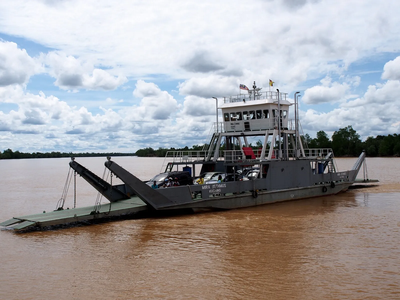 A grey vehicle ferry with a raised ramp crossing a wide brown river under a cloudy sky.