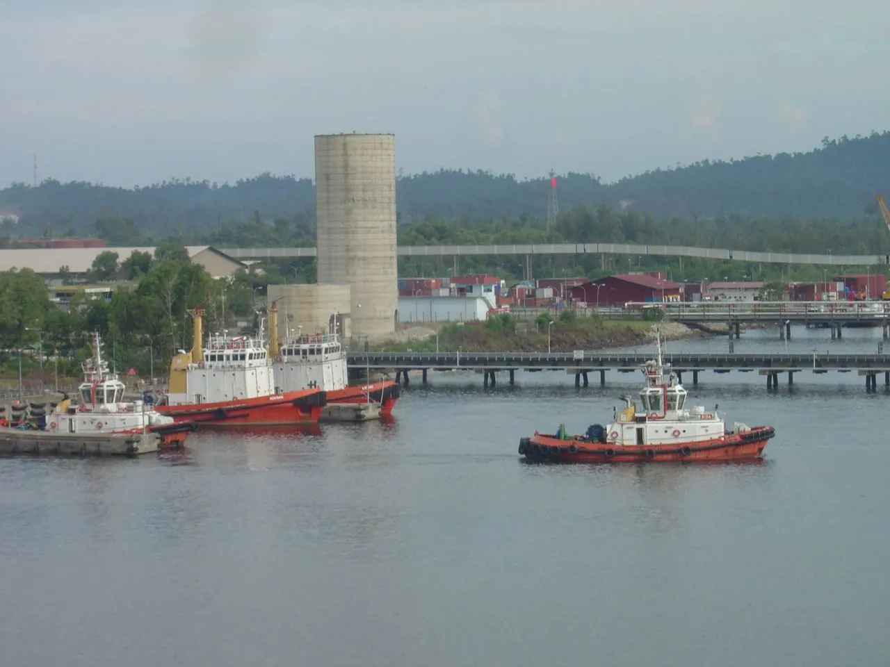Several red and white tugs and small boats moored at a jetty, with a tall concrete silo and green hills behind.