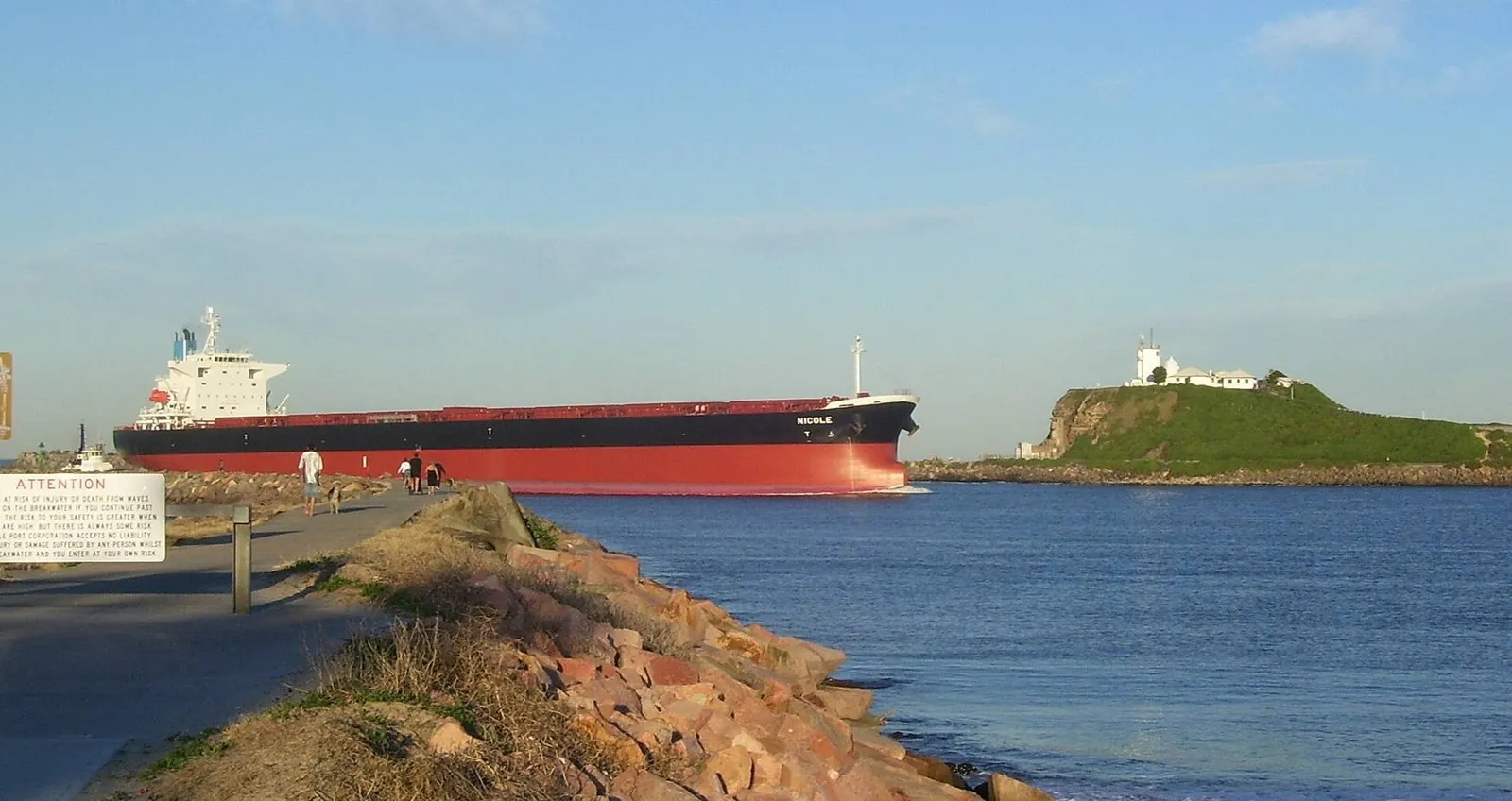 A bulk carrier with a red hull and white bridge sails past a grassy headland with a lighthouse, seen from a rock breakwater.