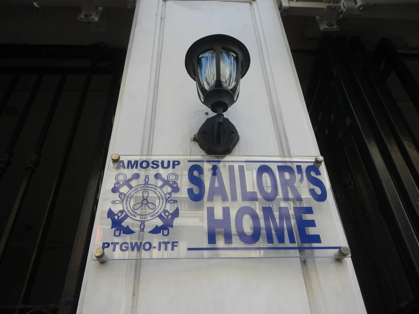 A clear sign on a white pillar under a black lamp. It shows the AMOSUP PTGWO-ITF union logo and the words Sailor's Home in blue.