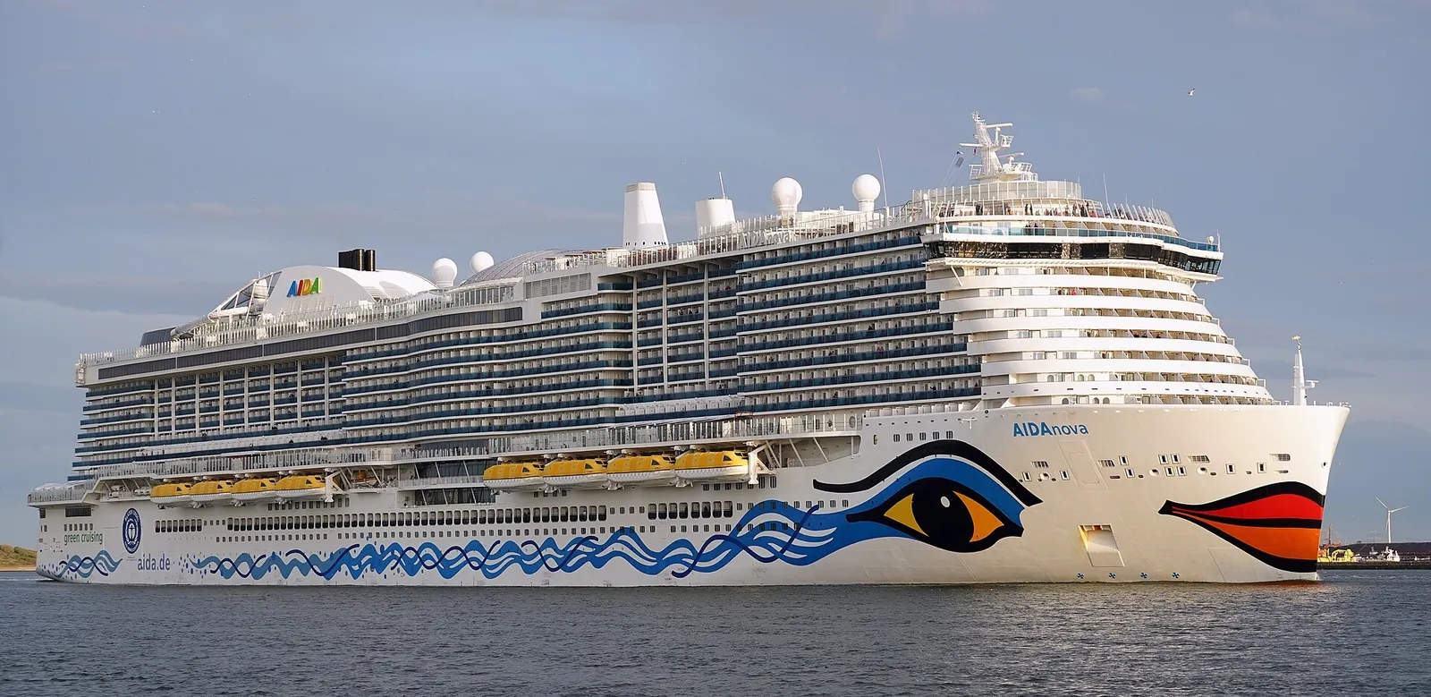 A large white cruise ship with a painted eye and red lips on its bow and a row of yellow lifeboats along its side, sailing in calm water.