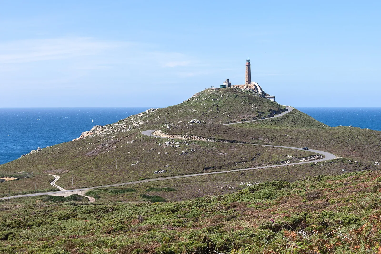 A stone lighthouse on top of a green hill above the sea, with a narrow road winding up to it.