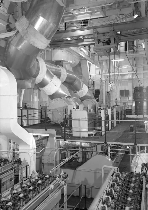 Black and white photo of a large ship engine room with pipes, walkways and a crane on beams near the ceiling.