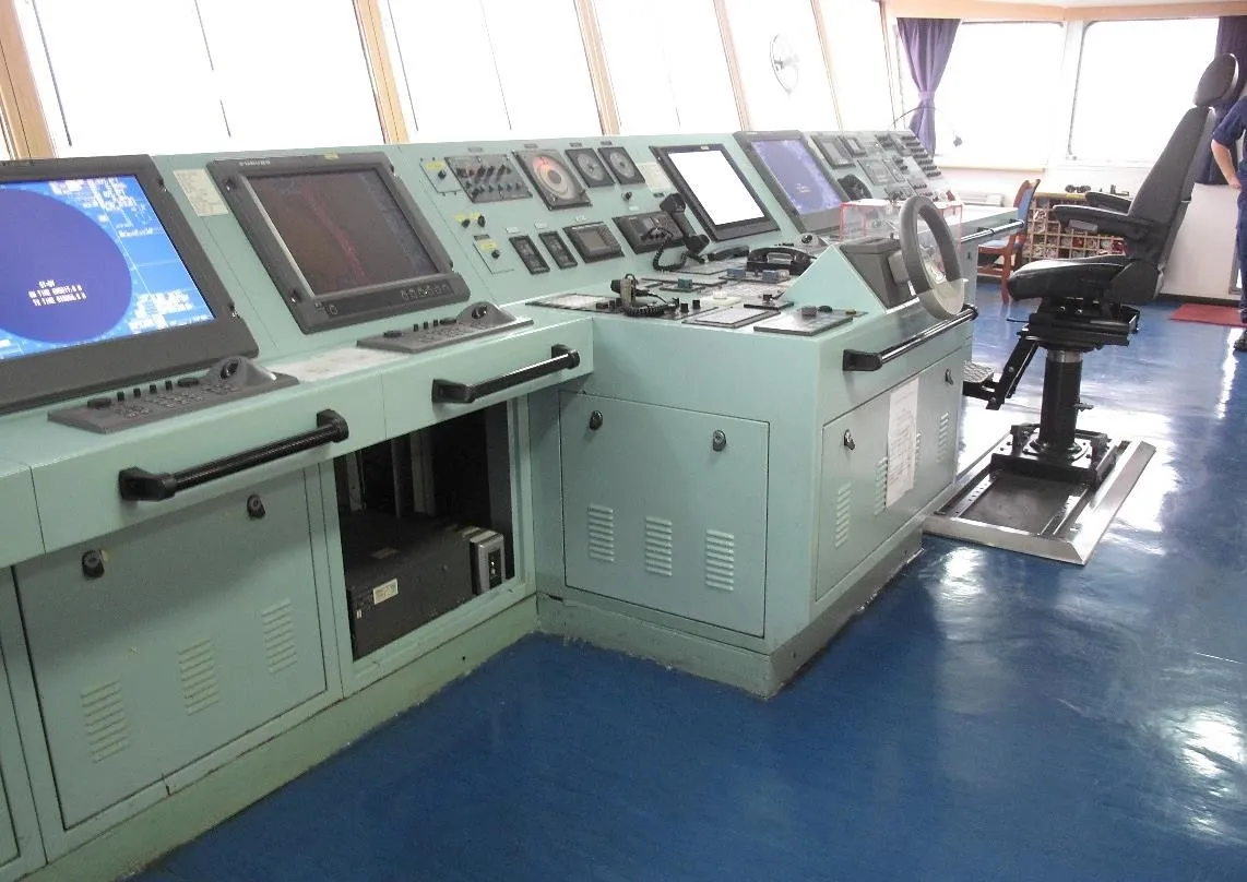 Bridge consoles of a merchant ship with radar and chart displays, a steering wheel and a pilot chair on a blue floor.