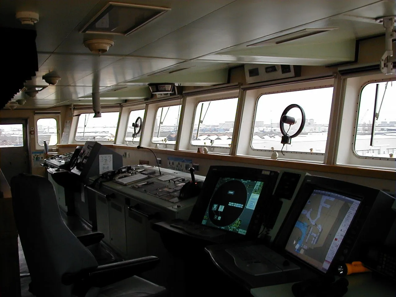 The empty bridge of a cargo ship with a radar screen, an electronic chart screen, a chair and large windows looking out at a port.