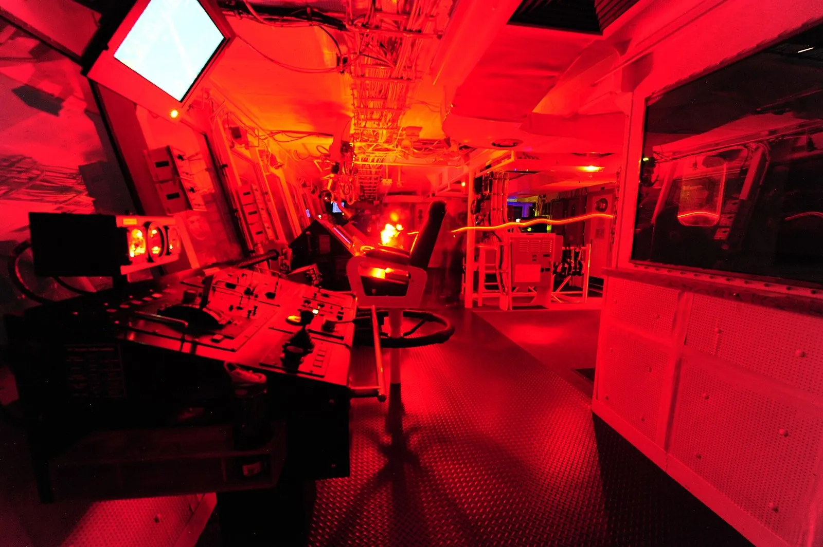 A ship's bridge at night lit by red light, with a control console, a chair and screens.