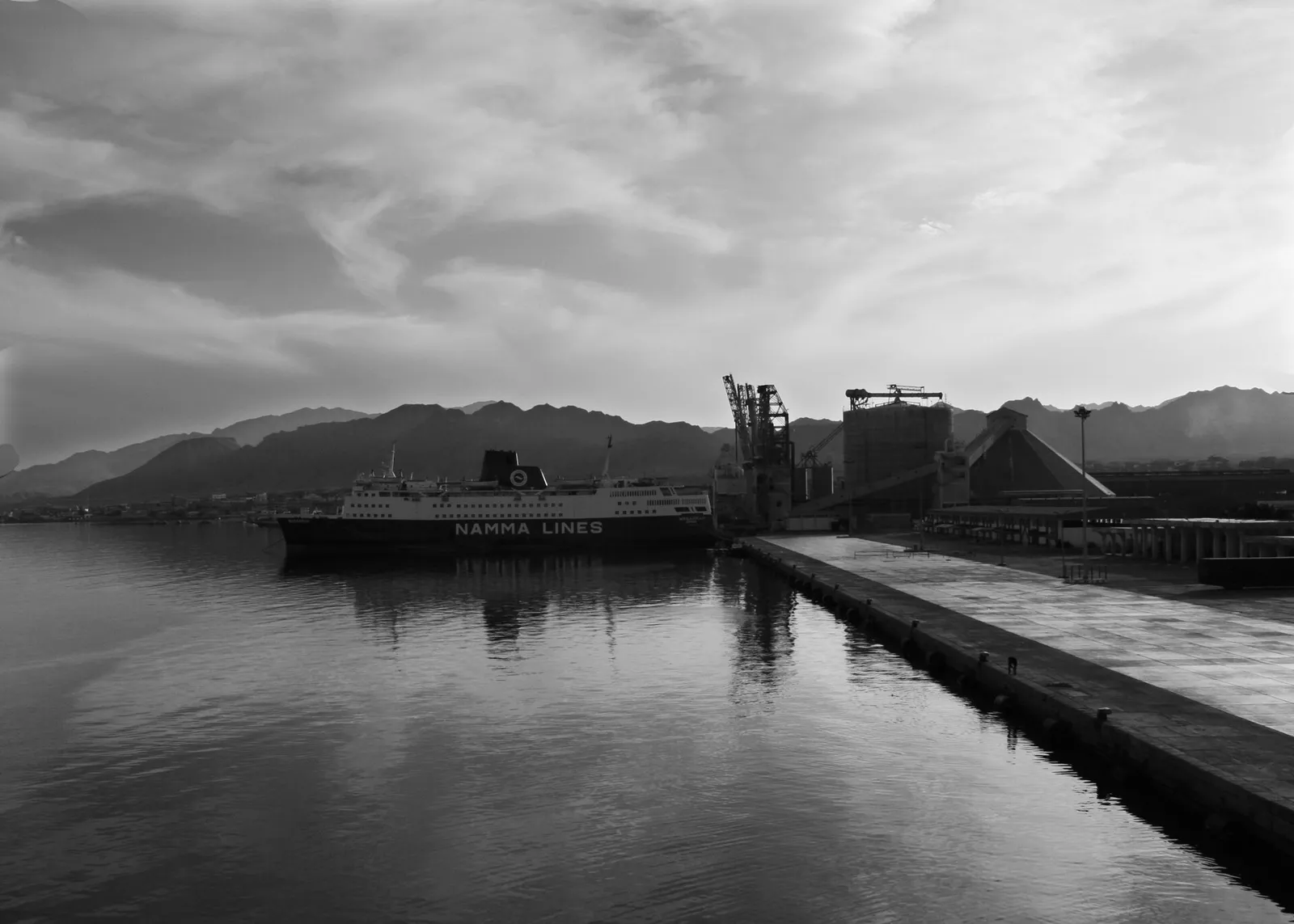 Black and white photo of a passenger ferry moored at a long concrete quay, with cranes, storage buildings and mountains behind it.