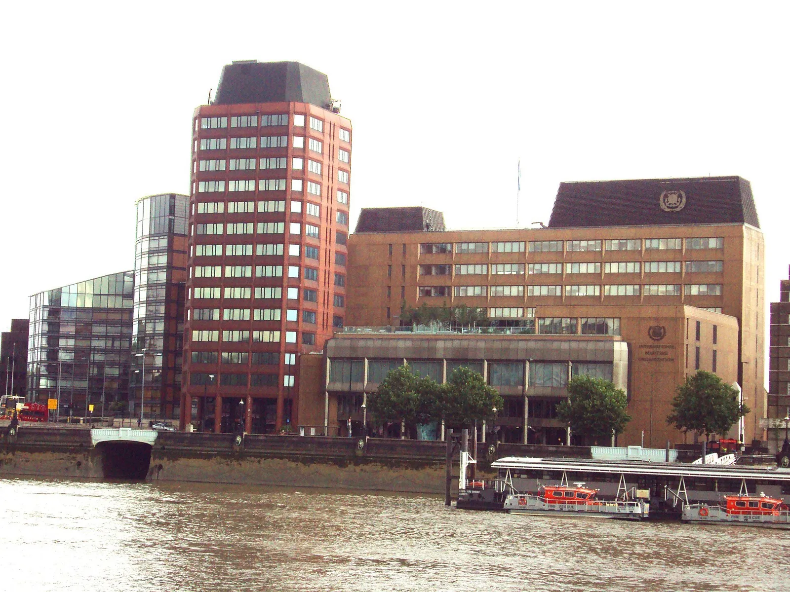 A large brown office building with the IMO emblem on its roof, beside the river, with a red tower block to its left and moored boats in front.